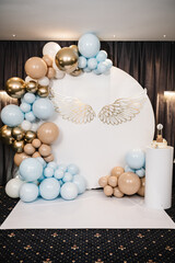 Celebration baptism concept. Birthday decor for a baby boy party on white background. Photo wall decoration of angel wings and white, blue, brown, and gold balloons in hall restaurant.  Trendy cake.