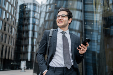 Optimistic businessman in suit using phone with reflective city buildings in the background, a...