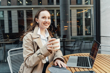 Smiling woman with coffee and laptop outdoors, enjoying a work break, city life merging with remote...