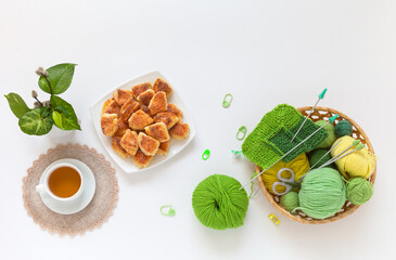 Top view of wicker basket with needlework and hand knitting and cup of tea and homemade fresh cookies on white table. Evening gatherings with tea and handicrafts. Flat lay, copy space, mock up