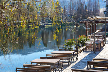 Fototapeta na wymiar Cafe on river bank. Springtime willows leaves at foreground, calm water, sky and trees reflected in. Shallow depth. Old Town (Stari Grad) of Trebinje, Bosnia and Herzegovina