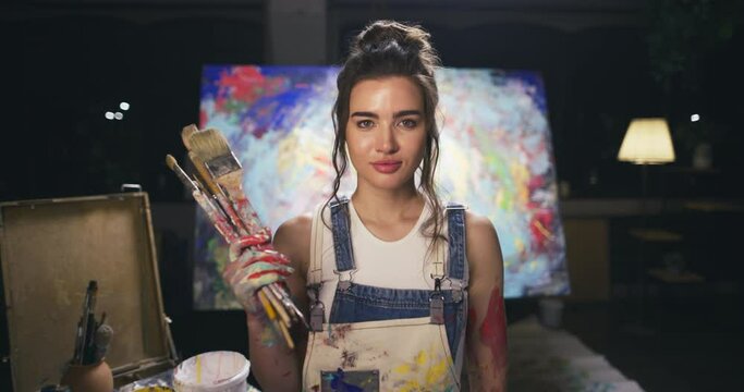 Portrait of talented female artist paint dirty with set brushes in her hands, wear apron abstract modern art paint picture on background. Concept of creative profession, inspiration education drawing 