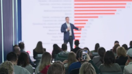 Pan right real time of male influencer standing on stage and explaining business ideas to crowd against big screen with statistics