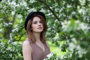 Fototapeta premium Stylish young adult woman female model with clean fresh skin, make-up and brown hair in blossom garden on green leaves background outdoor portrait