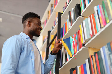 A serious, intelligent black student stands in a library, carefully selecting books from the bookshelves for research.