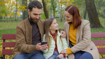 Family together happy man father woman mother child kid girl little daughter outdoors park on bench dad mom baby using mobile phone smartphone browsing choosing goods shopping online talking in city