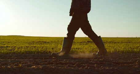 Farmer walks in rubber boots  down a farmer field  dust rising from shoes. Low angle. One part is sown, the second part is not sown.
