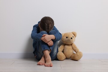 Child abuse. Upset girl with toy sitting on floor near white wall