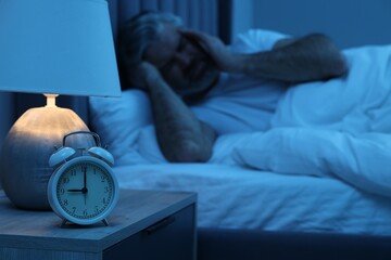 Mature man suffering from headache in bed at night, focus on alarm clock