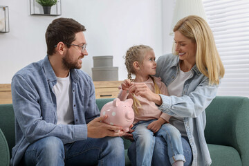 Planning budget together. Little girl with her parents putting coins into piggybank at home
