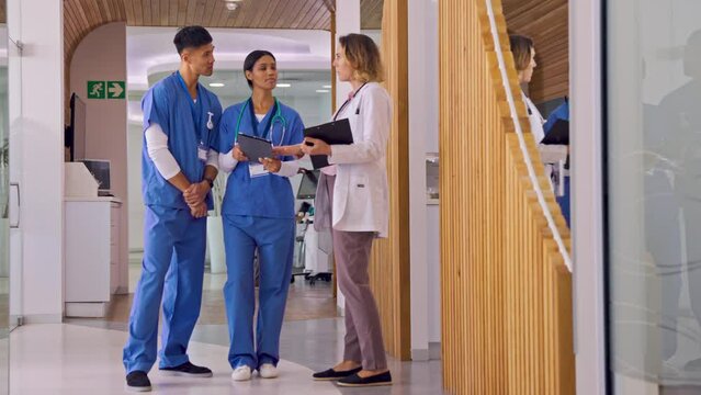 Doctor Talking With Nurses As They Walk Along Corridor In Modern Hospital With Digital Tablet