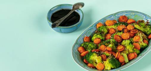 Asian-style stir-fried broccoli with carrot. Dark sauce in a small bowl with a spoon lying on it....