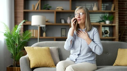 Senior gray haired female talking on phone while sitting on sofa in living room at home. Elderly woman has a conversation with a colleague or communicates with a friend or family using a smartphone