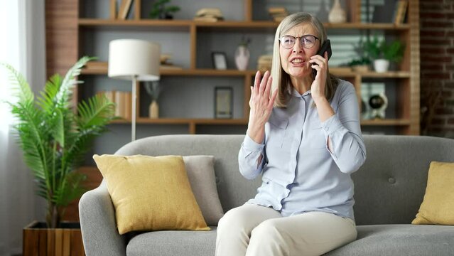 Angry disappointed senior female talking arguing over phone sitting on sofa in room at home. Frustrated elderly woman with gray hair complains about problems that need to be solved using a smartphone