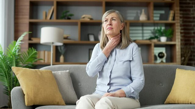 Sck senior gray haired female suffering from a sore throat sitting on sofa in living room at home. Elderly retired woman has symptoms of a cold, virus or flu. She feels pain and massaging a sore spot