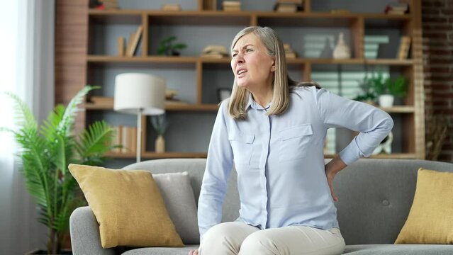 Tired senior gray haired female suffering from back pain sitting on sofa in living room at home. Upset elderly retired woman massages painful lower back muscles. She is exhausted or has sciatica