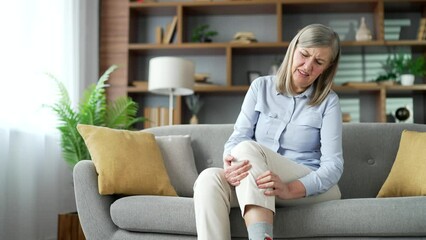 Senior gray haired female suffers from leg pain, calf muscle spasm sitting on sofa in living room at home. Elderly woman massaging leg calf muscle cramps, trauma from inflammation of tendon at calves