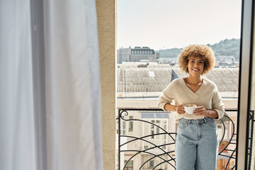 Fototapeta na wymiar smiling curly african american woman standing with cup of coffee on hotel balcony, city view