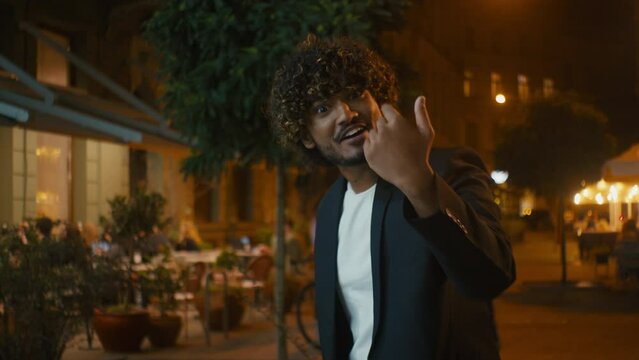 Young Indian Arabian Latino American man calling inviting hand gesturing city street outside evening enjoy come here welcome friendly smile approval offer propose sign joyful join excited playful guy