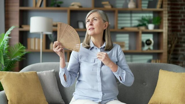 Senior gray haired female is hot while sitting on sofa in the living room at home. Elderly retired woman waves a fan, feels discomfort and sweating, because the air conditioner in apartment is broken
