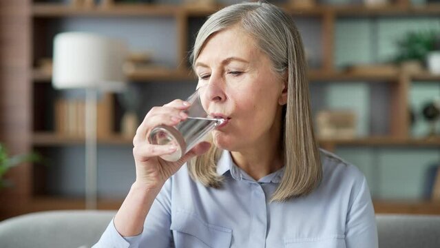 Senior female drinks water from a glass while sitting on the sofa in the living room at home. Happy smiling elderly woman feeling relieved, enjoying a clean cool drink, relaxing and resting. Close up