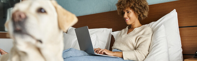 young african american woman working on laptop with her labrador on bed in a hotel room, banner