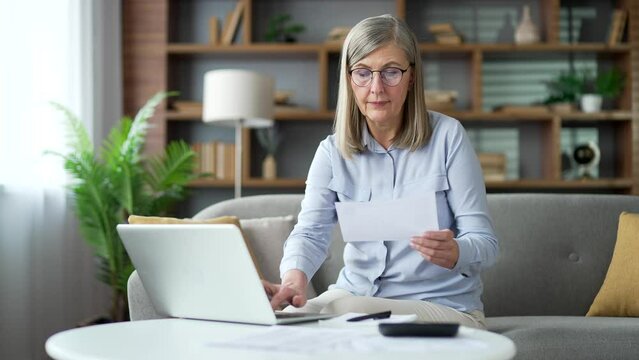 Happy senior gray haired female uses application for spending bills using laptop sitting at home office. Elderly woman calculates household finances, enters data into a chat bot easy and simple to pay