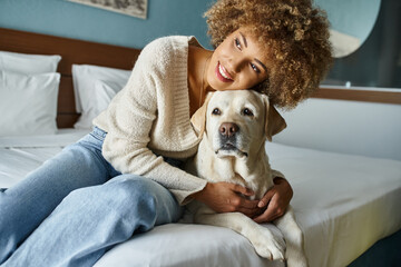 curly happy african american woman smiling while cuddling labrador in pet-friendly hotel room