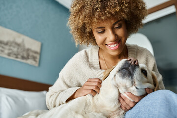 cheerful young african american woman cuddling labrador on a bed in a pet-friendly hotel room