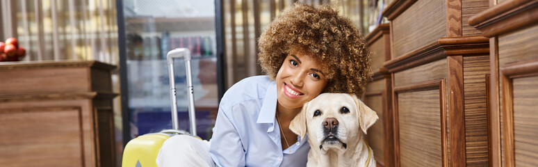 banner of happy african american woman cuddling dog near suitcase in lobby of pet-friendly hotel