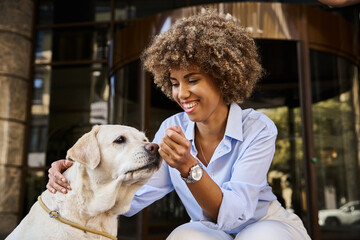 Smiling and curly african american woman cuddling labrador outside a pet-friendly hotel