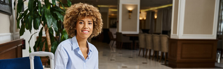 happy african american woman with curly hair waiting for check in hotel lobby, horizontal banner