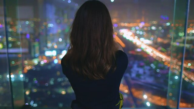 Woman make panoramic shot of night big city lights. Aerial cityscape view from top floor skyscraper window. Colorful bokeh in background. Girl take photo of urban traffic on mobile phone, back view