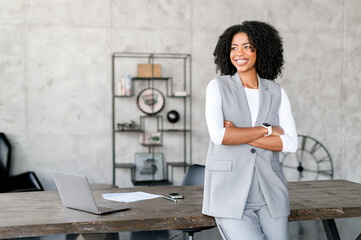 A joyful African-American businesswoman leaning on a desk in office space, her attire and demeanor...