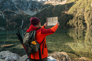 Shot of lost woman traveler with map and backpack relaxing outdoor with rocky on Morskie Oko lake. Woman traveler explorer with backpack hiking to the top of the mountain. Hiking travel and adventure - obrazy, fototapety, plakaty