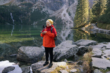 Rear shot of traveler 60s woman influencer in sportswear with backpack standing on the rocky shore and making selfie on smart phone. Taking photo of green hills and mountains on Morskie Oko lake - obrazy, fototapety, plakaty