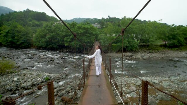 A woman in white clothes walks backwards along a suspension bridge over a mountain river.