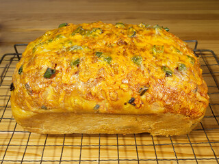 Closeup of an oven fresh, uncut loaf of baked homemade bread on a cooling grid, made with chopped green jalapeno peppers and a melted cheese topping.