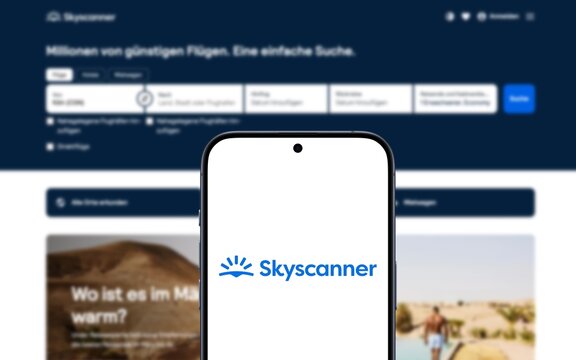 Skyscanner-Logo wird auf einem modernen Smartphone angezeigt, Website des Suchaggregators und Reiseb&uuml;ro im Hintergrund, Reiseoptionen recherchieren und buchen, einschlie&szlig;lich Fl&uuml;ge, Hotels 