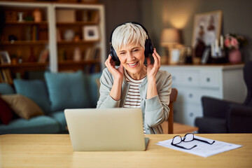 A delighted senior adult woman with headphones listening to a web conference from home on a laptop