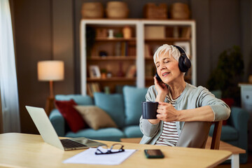 A relaxed senior adult woman with headphones listening to music and singing while having a cup of coffee and sitting in front of a laptop