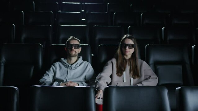 A couple in a movie theater is watching a 3D movie in a cinema. A woman drinks soda from a red cup