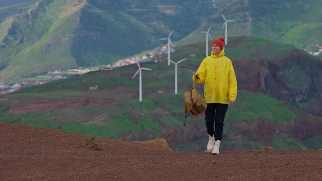Attractive traveling woman in a yellow jacket and backpack looking at windmills in field 