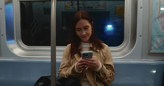 Young stylish brunette female using smartphone texting and smiling taking the subway