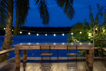 Obraz premium Along the waterfront, there are dining tables, chairs, and beautifully decorated lights that shine brightly during Twilight, ready for dining in a luxury restaurant. There are shady trees around.