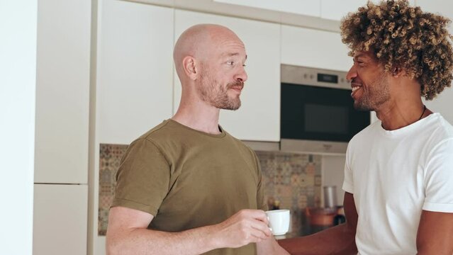 A loving gay couple shares a moment of happiness in the kitchen. They are smiling and engaged in a relaxed morning conversation.