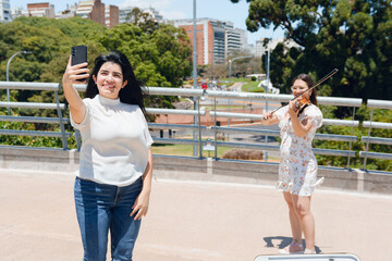 happy latin woman taking selfie with her phone with violinist busker woman in background