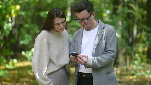 A man and woman in formal wear stand in the woods, sunglasses on, looking at a cell phone. Green grass, trees surround them as they gesture at the screen