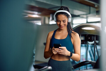 Happy athletic woman listening music over headphones while using mobile phone during gym workout.