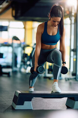 Young sportswoman with dumbbells exercising on step aerobics equipment in gym. - obrazy, fototapety, plakaty